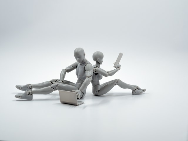 two grey androids sitting on the ground. one with a laptop and one with a tablet